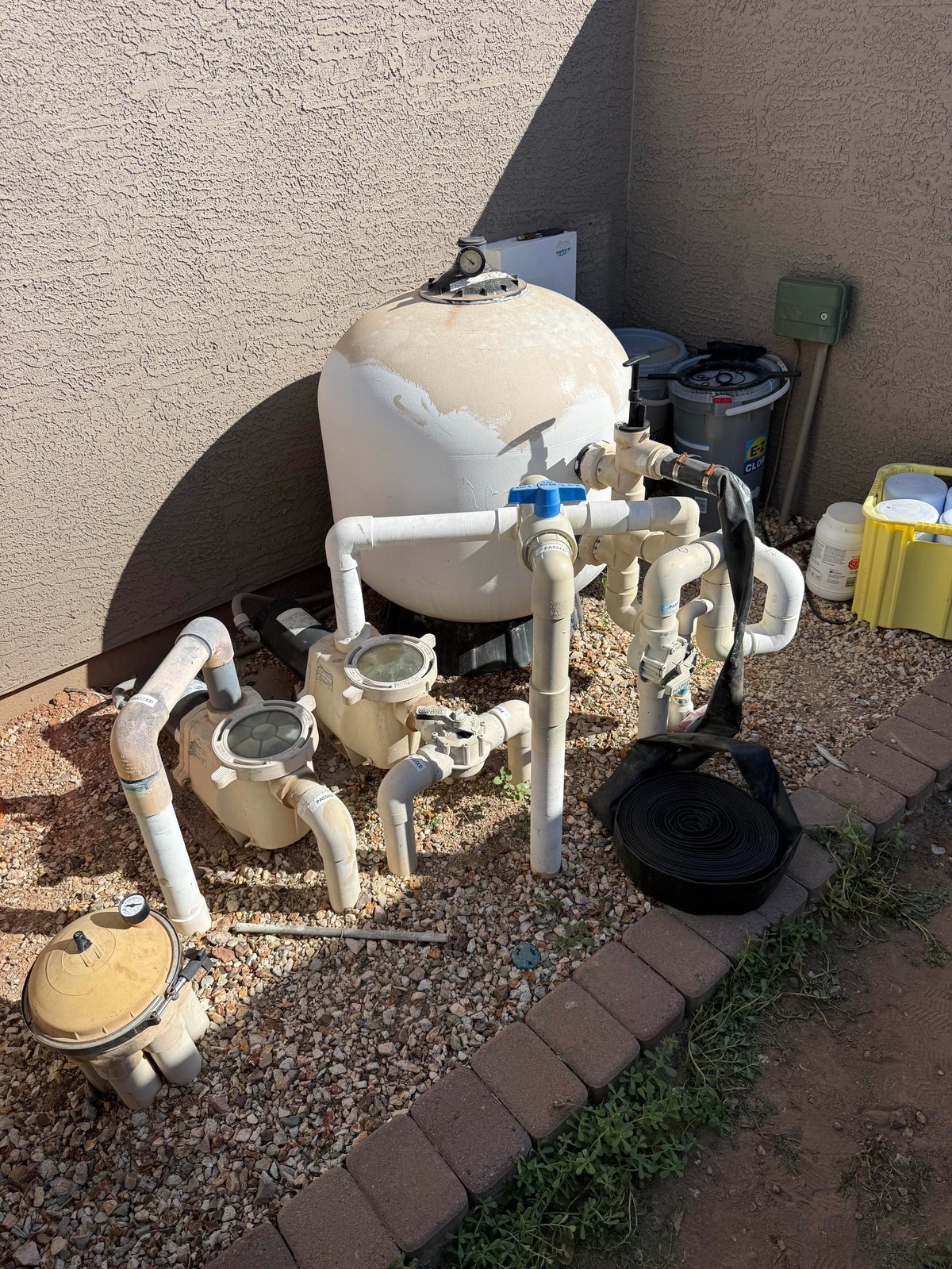 Outdoor water filtration system with white tanks and PVC pipes against a concrete wall next to gravel and brick edging