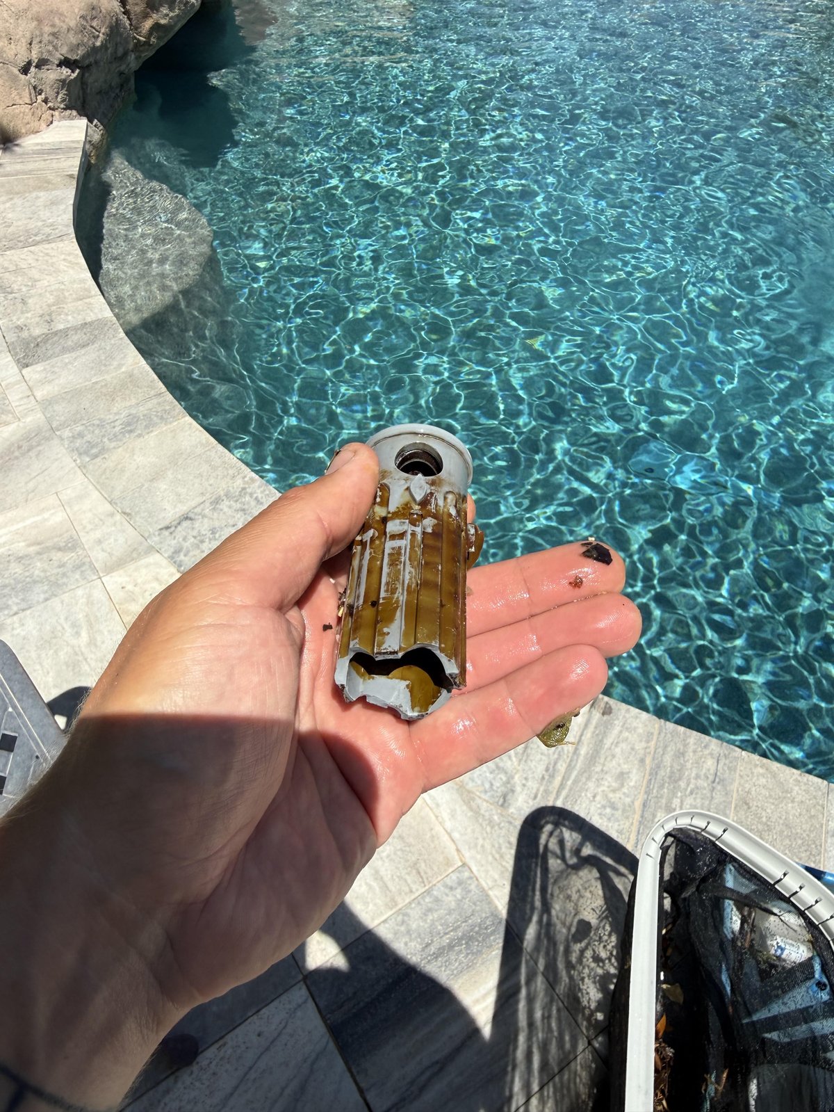 Hand holding a corroded brass object over a clear turquoise swimming pool by a stone deck