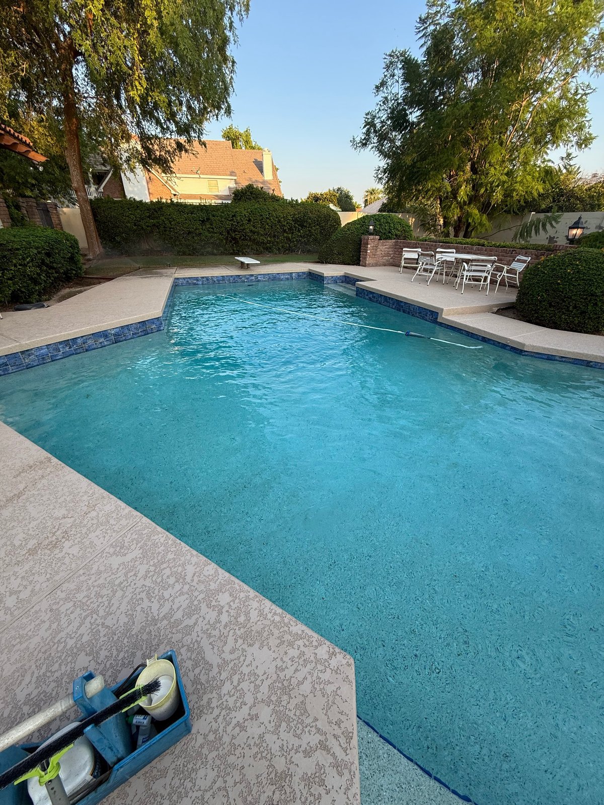 Backyard swimming pool with turquoise water, concrete deck, mature trees, hedges, and patio seating area on a sunny day