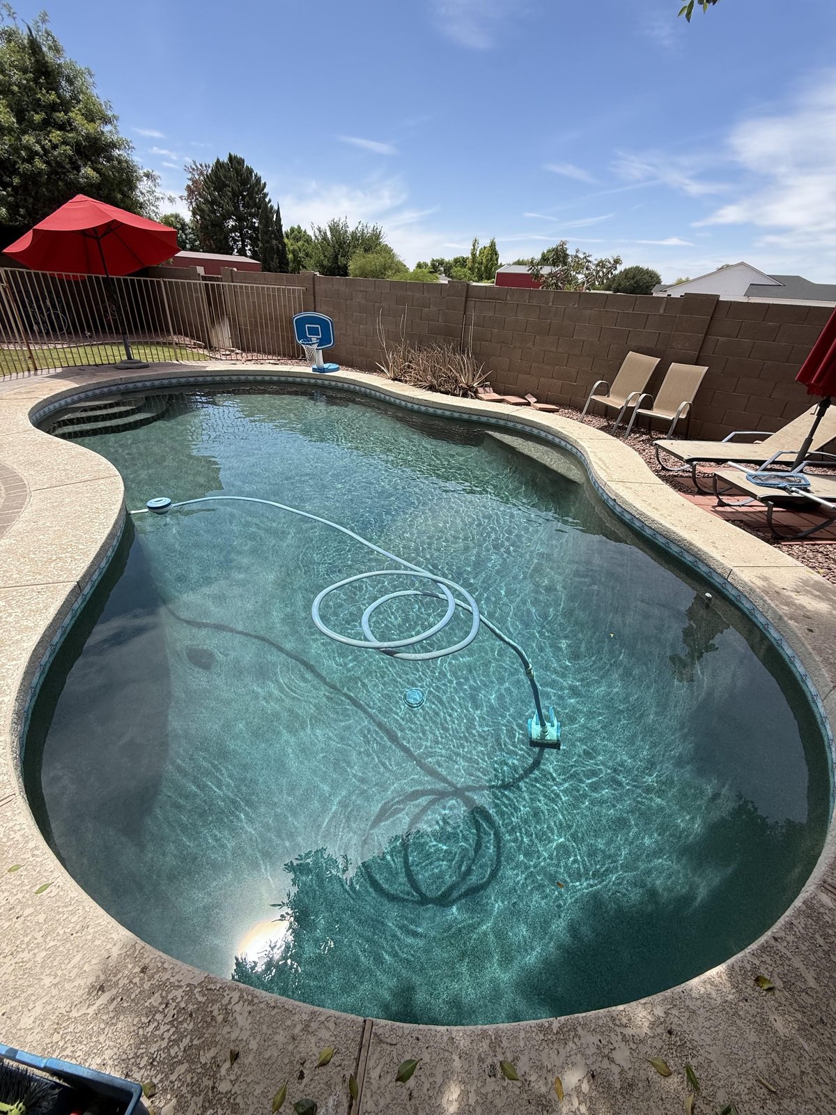 Recovered kidney-shaped backyard pool with clear turquoise water, pool cleaning equipment, lounge chairs, and red umbrella