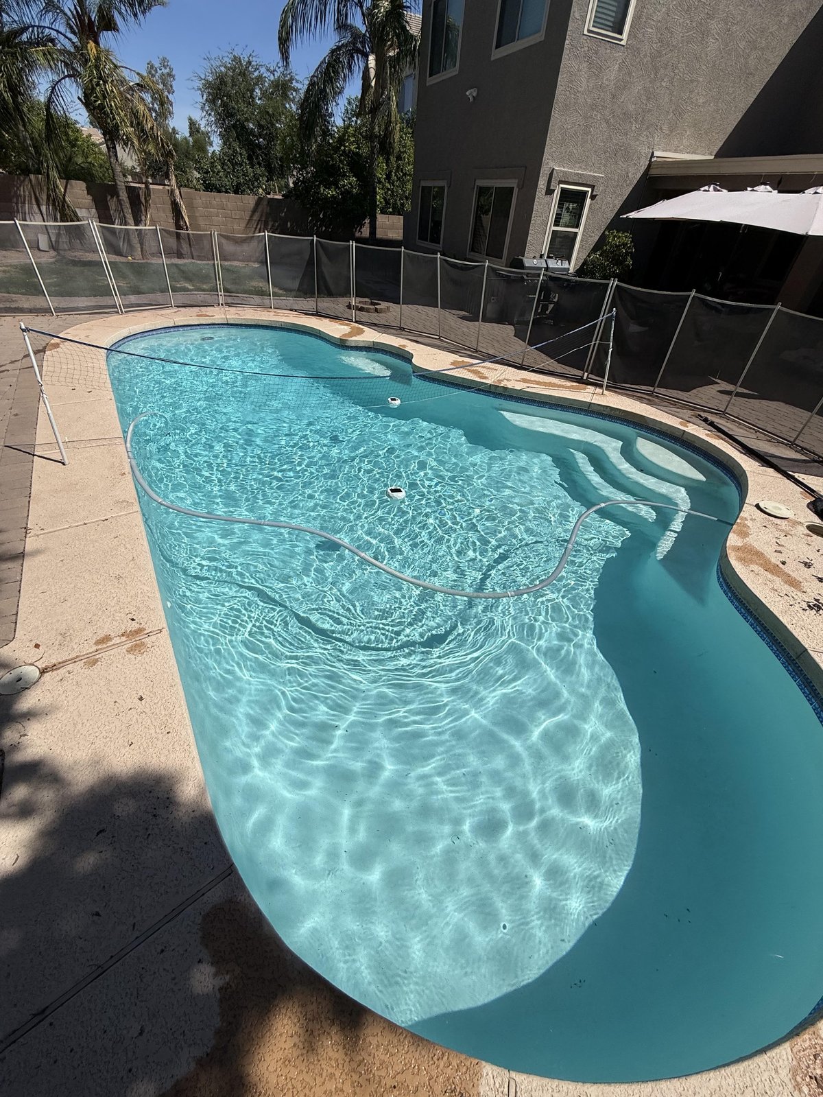 Bright turquoise swimming pool with clear water and ripples, surrounded by tan concrete deck, black safety fence, dark building, and palm trees.