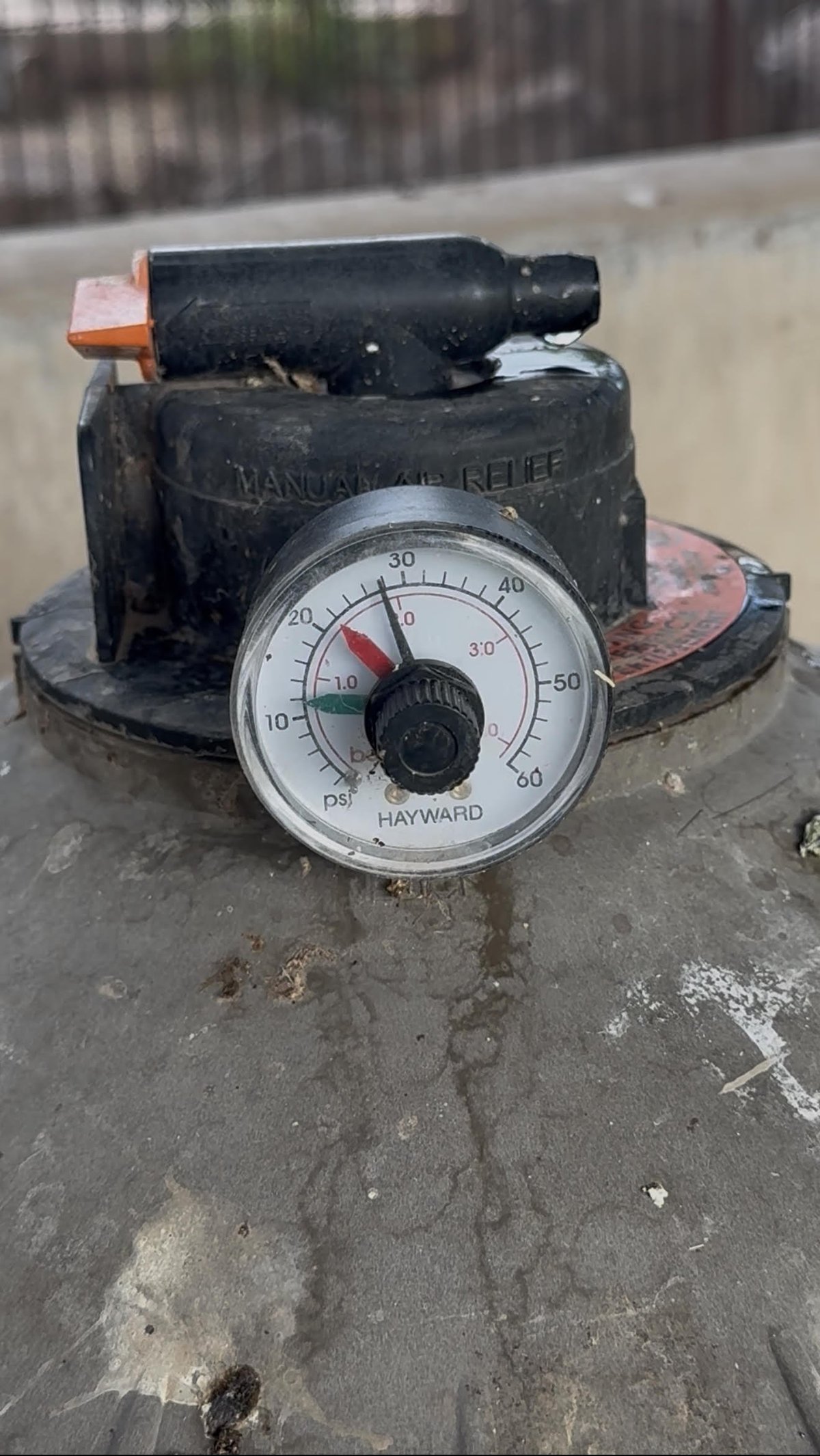 Weathered Hayward pool pump pressure gauge sitting on concrete surface with blurred fence background