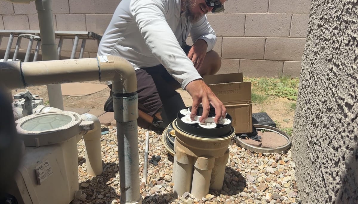 Person in white shirt performing maintenance on outdoor plumbing equipment near a brick wall