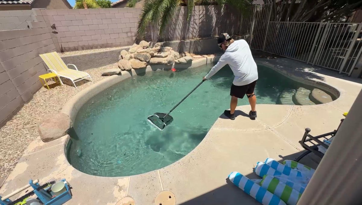 Man cleaning backyard pool with net skimmer on sunny day